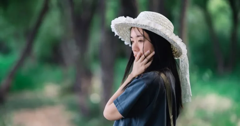 A thoughtful young woman in a straw hat enjoys a serene moment in a lush green forest setting.