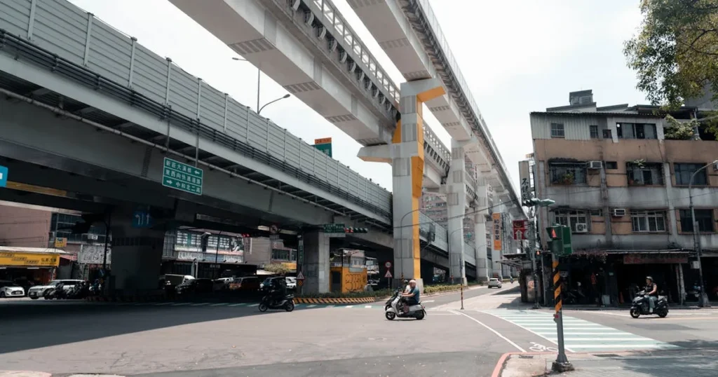 Motorbikes navigate an urban intersection beneath towering elevated train tracks in bustling New Tai