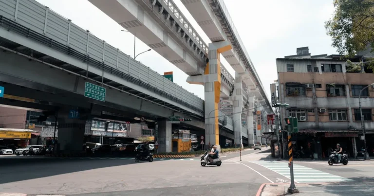 Motorbikes navigate an urban intersection beneath towering elevated train tracks in bustling New Tai