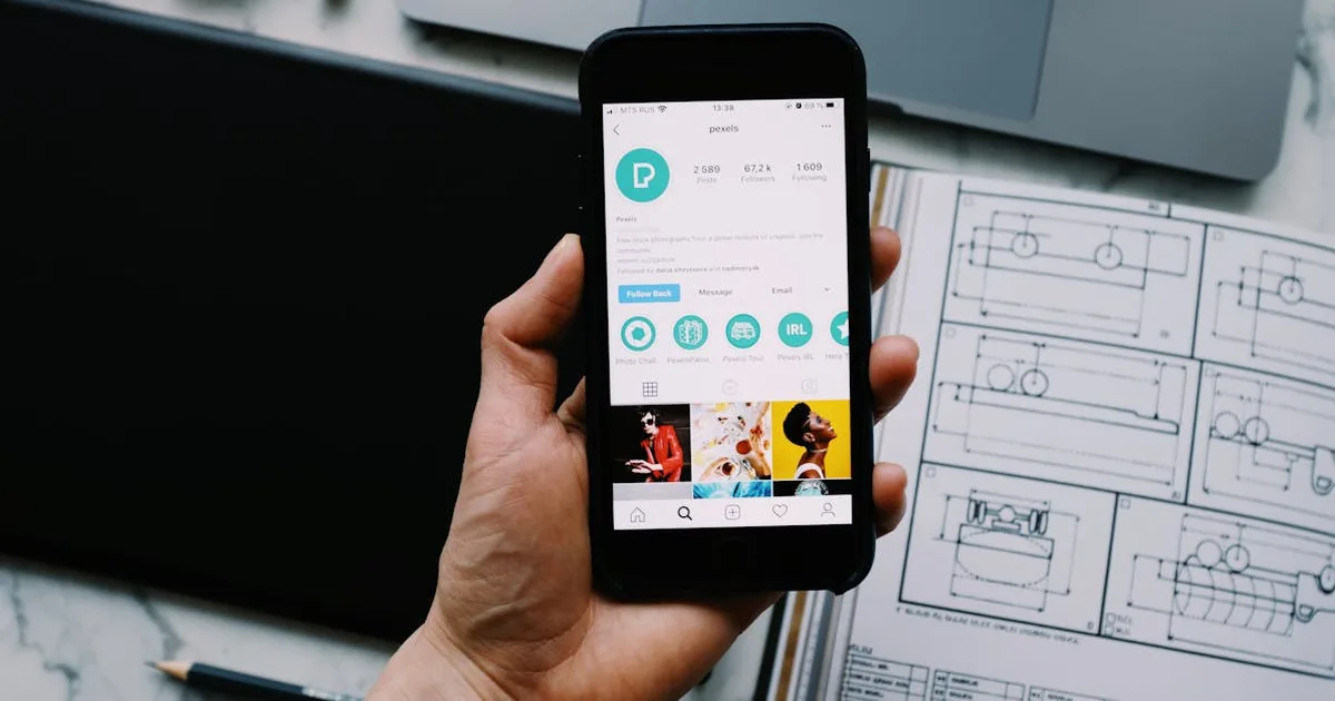 Hand holding smartphone displaying social media profile with tech gadgets on desk.