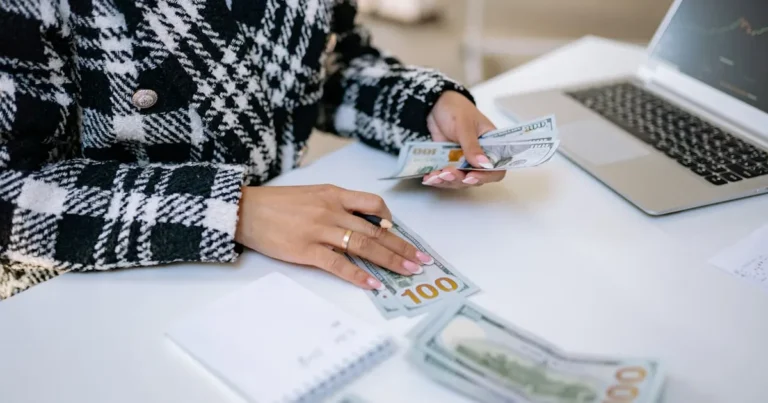 Close-up of a woman counting hundred-dollar bills at a desk with a laptop, focusing on finance and w